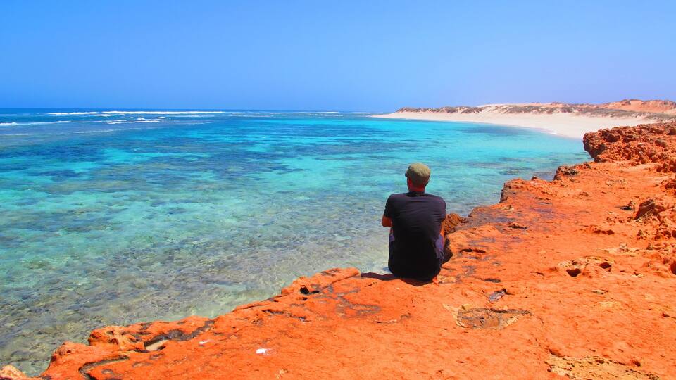 A man sitting on a red rock overlooking a clear blue sea and a sandy beach.