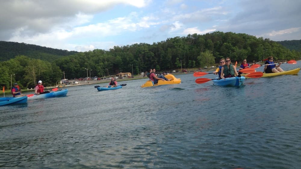A group of people kayaking on a calm lake with a forested hill in the background.