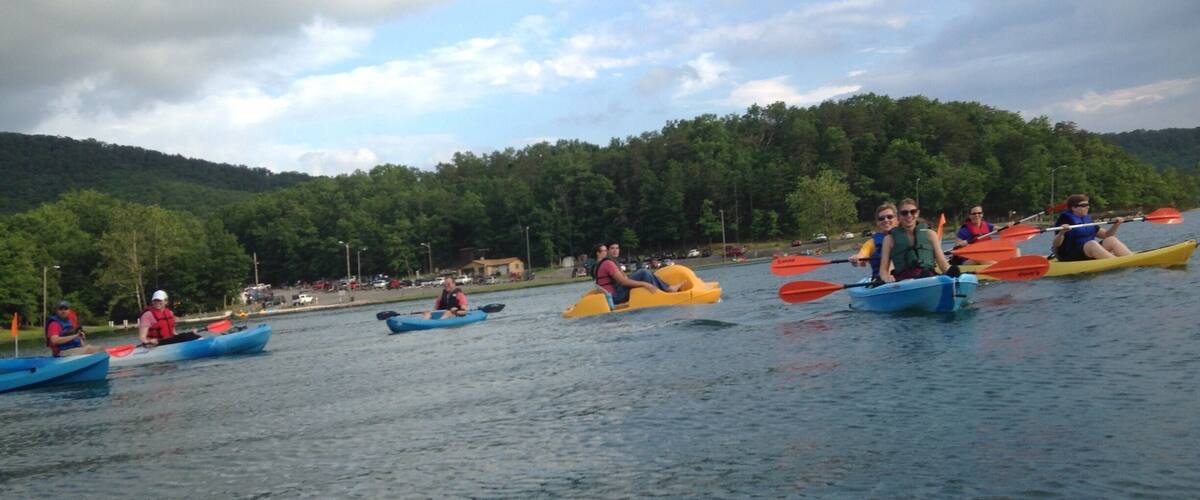 A group of people kayaking on a calm lake with a forested hill in the background.