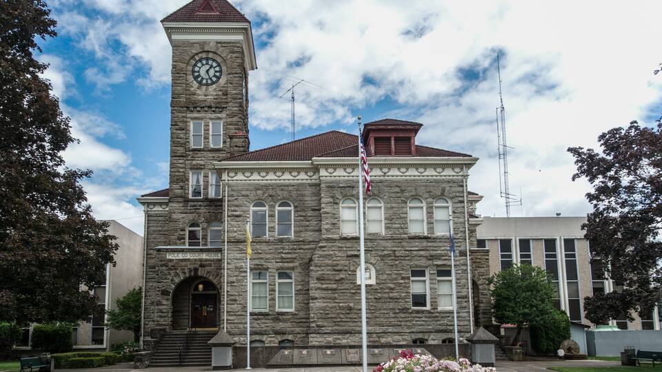 A historic stone courthouse with a clock tower, flagpole, and a well-maintained lawn.