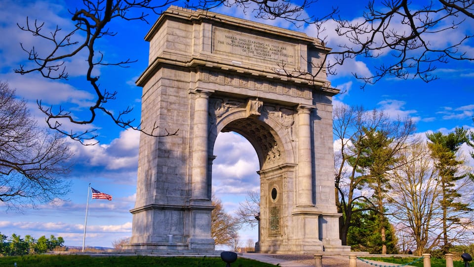 The National Memorial Arch stands with the American Flag blowing in the wind to the left at Valley Forge National Park in Pennsylvania.