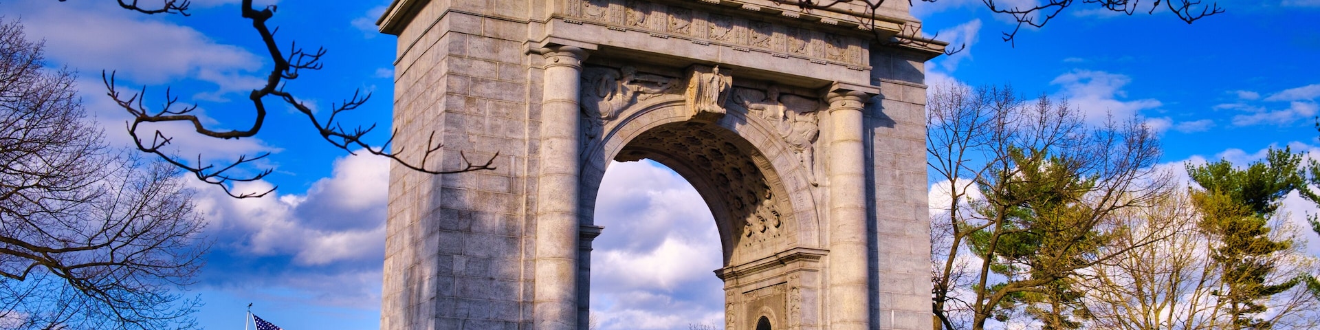 Un gran monumento de piedra en forma de arco con una bandera estadounidense, rodeado de árboles y un cielo despejado.