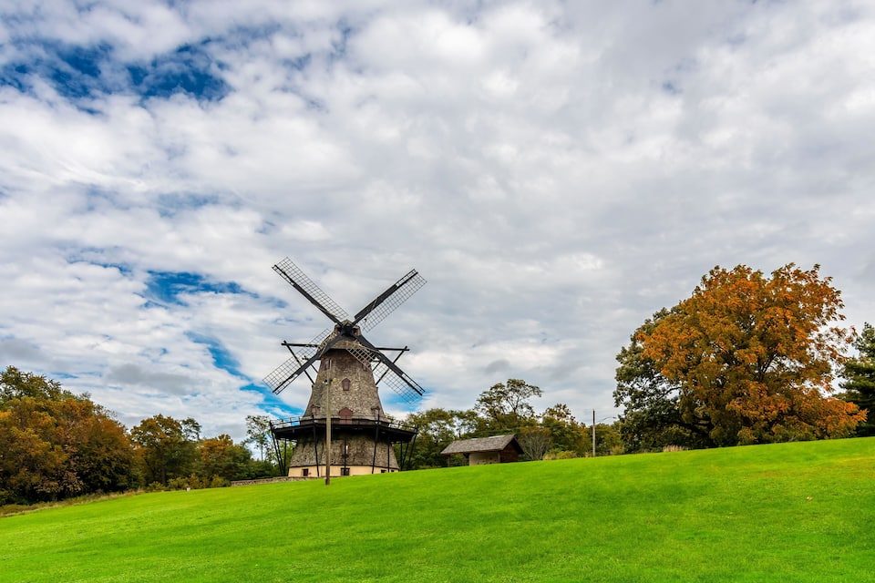 Een windmolen op een grasheuvel met herfstkleurige bomen en een gedeeltelijk bewolkte lucht.