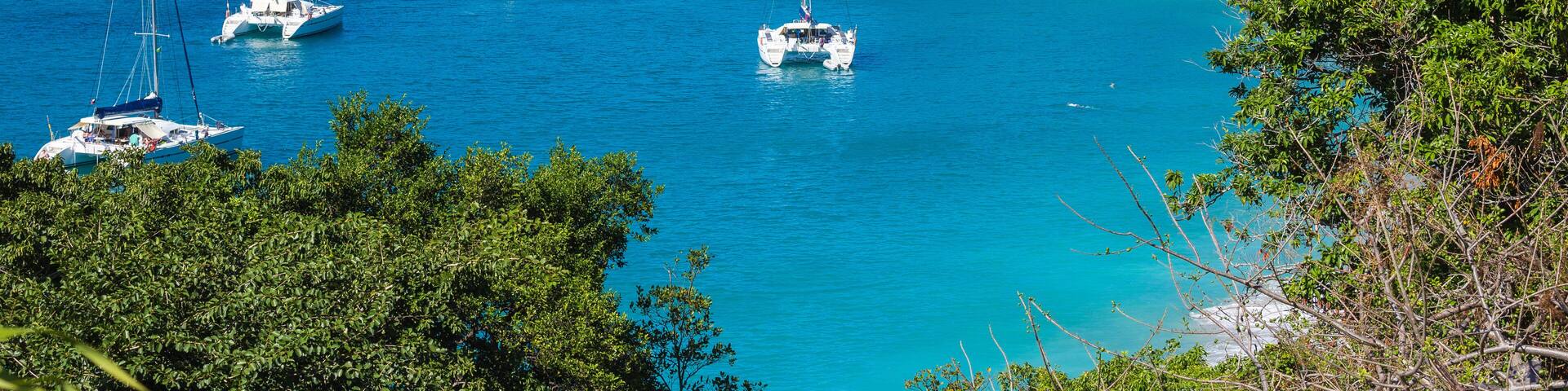 A coastal area with numerous sailboats anchored in clear blue waters, surrounded by lush greenery and hills in the background.