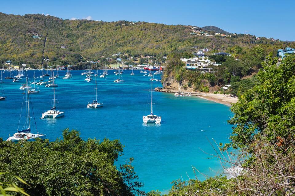A coastal area with numerous sailboats anchored in clear blue waters, surrounded by lush greenery and hills in the background.