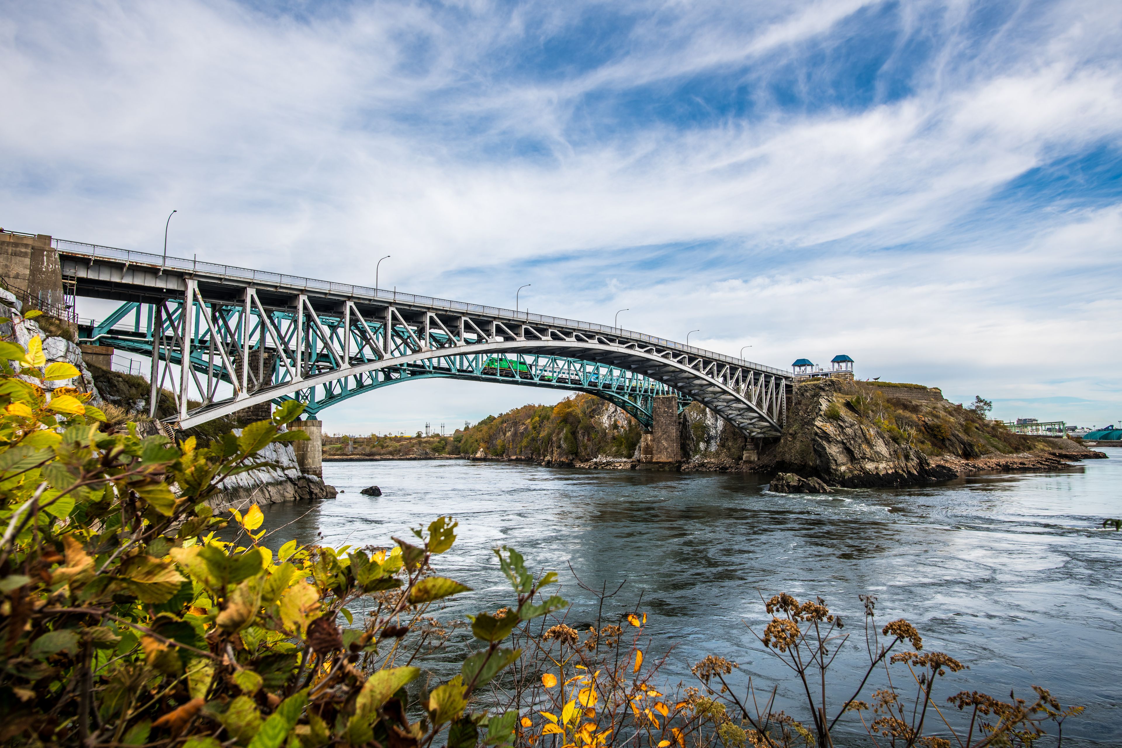 Un pont en acier aux couleurs verte et blanche enjambe une rivière, avec des affleurements rocheux de chaque côté et un ciel dégagé au-dessus.