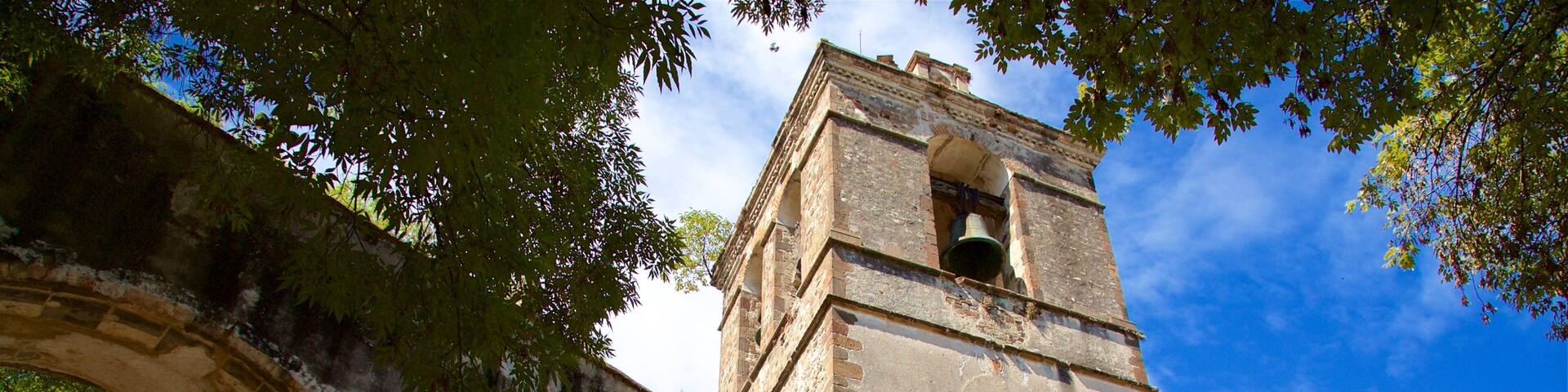 A historic stone tower with a bell, surrounded by an archway and trees.
