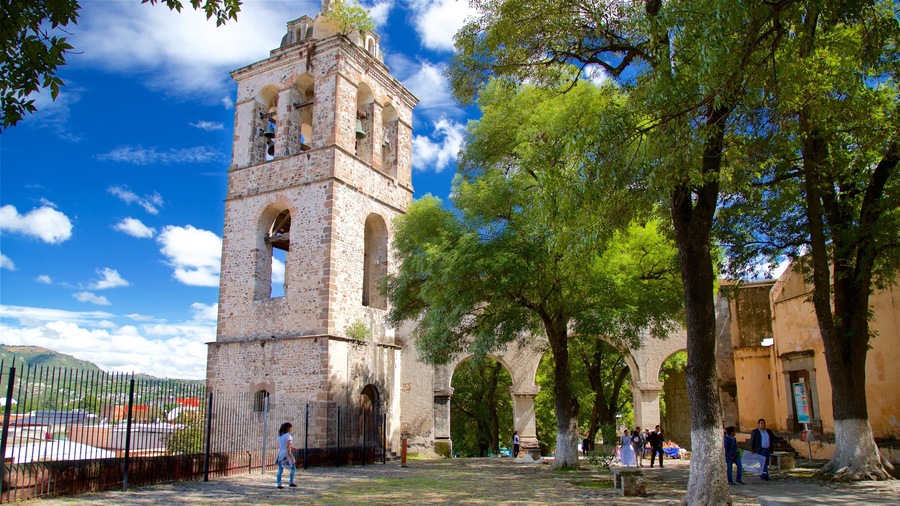 A historic stone tower with arched windows and a bell, surrounded by trees and a fence.