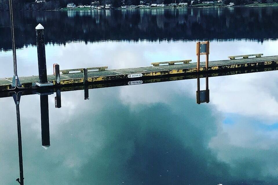Um lago calmo com um cais que se estende pela água, rodeado por uma encosta arborizada.