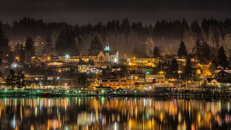 A nighttime view of a town with a church, houses, and a calm lake reflecting lights.