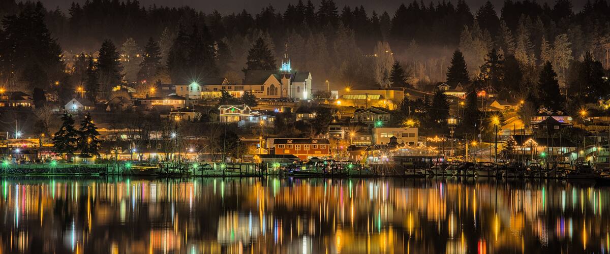 A nighttime view of a town with a church, houses, and a calm lake reflecting lights.