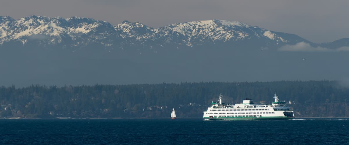 A ferry in a body of water with a forested coastline and snow-capped mountains in the background.