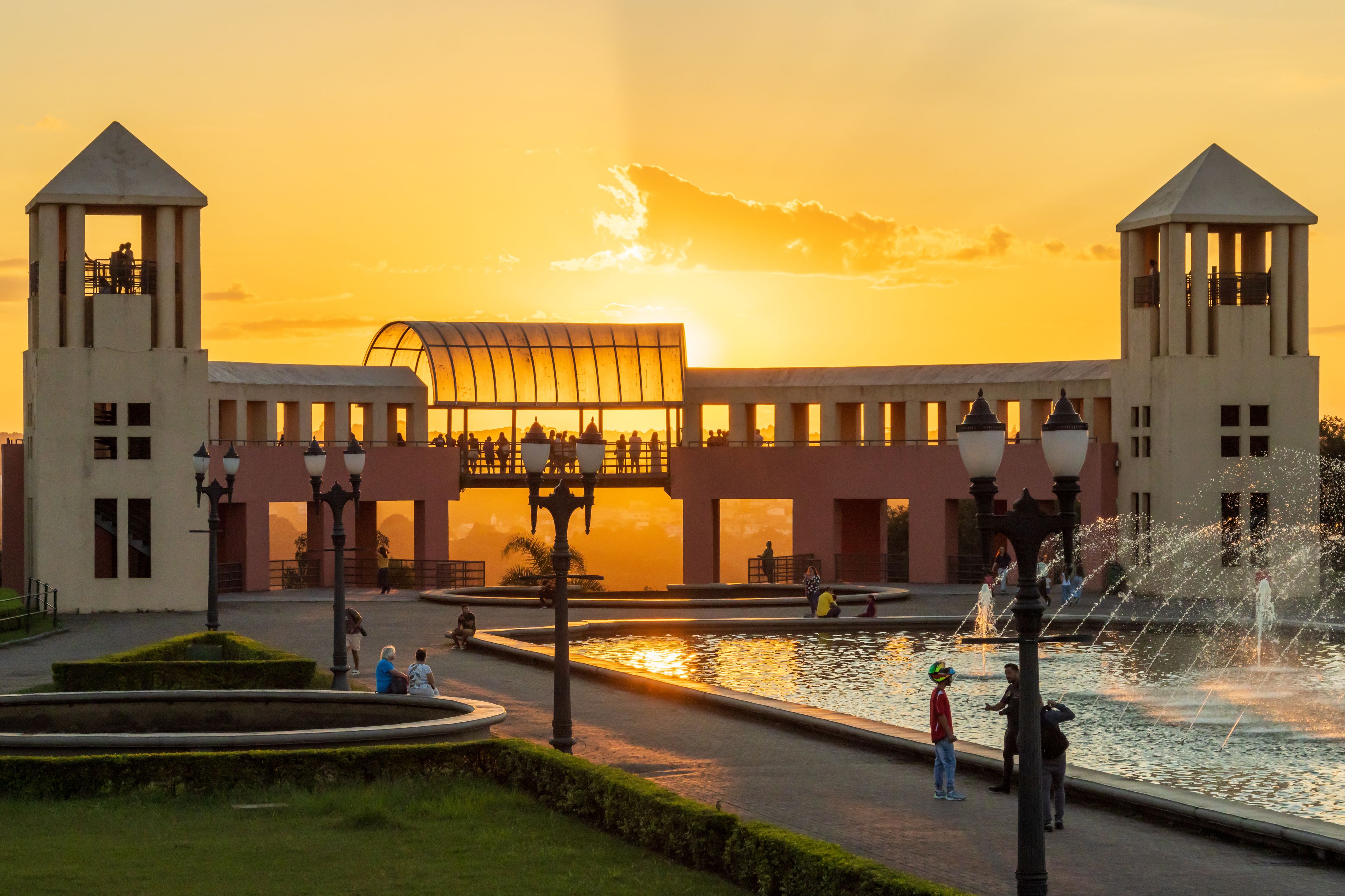 A sunset at a public square with a fountain, a glass-roofed building, and two towers.