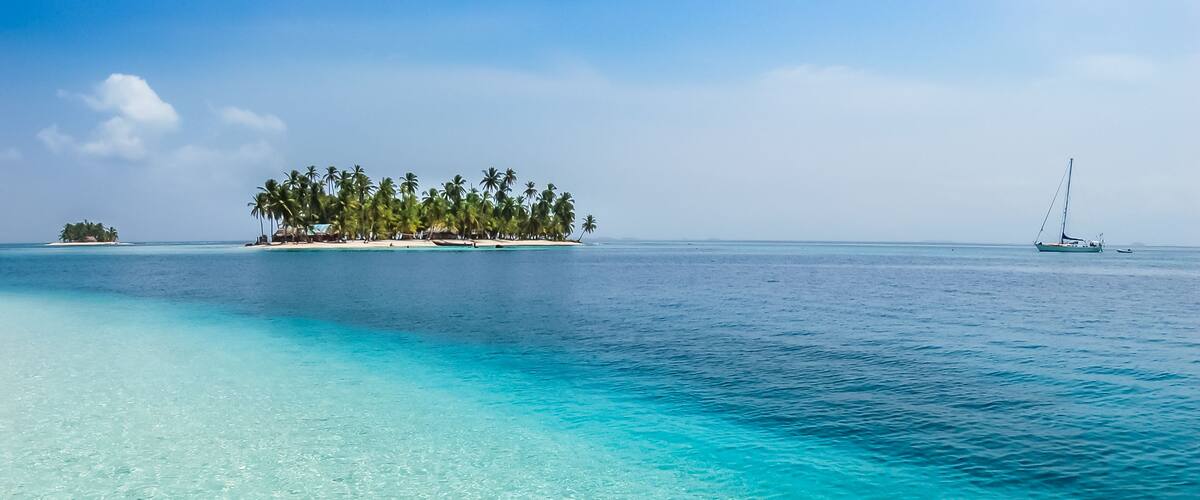 Un mar turquesa cristalino con una playa arenosa, una pequeña isla con palmeras y un velero a lo lejos.