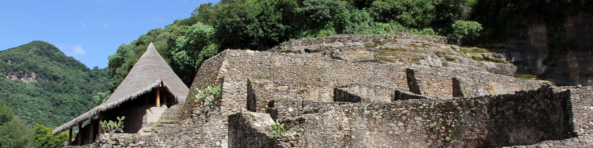 Antiguas ruinas de piedra con una estructura de techo de paja, en medio de un exuberante follaje y un paisaje montañoso.