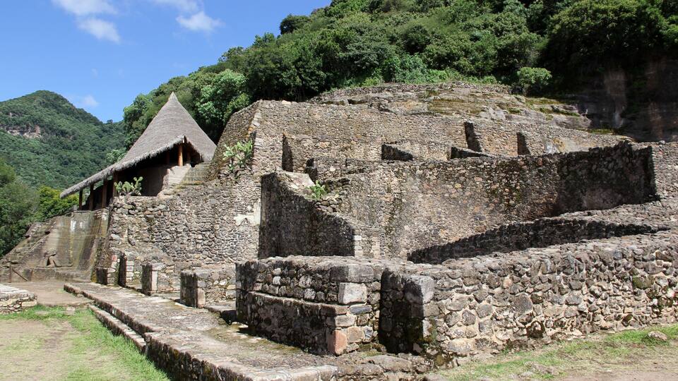 Ancient stone ruins with a thatched-roof structure amidst lush greenery and a mountainous backdrop.