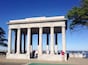 A neoclassical monument with columns, located near a waterfront with boats and people.