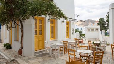A white building with yellow doors and windows, outdoor seating with white and wooden chairs, and a tree in the foreground.