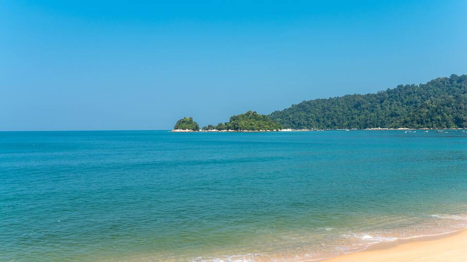 A beach with clear turquoise water, a sandy shore, and a forested hill in the background.