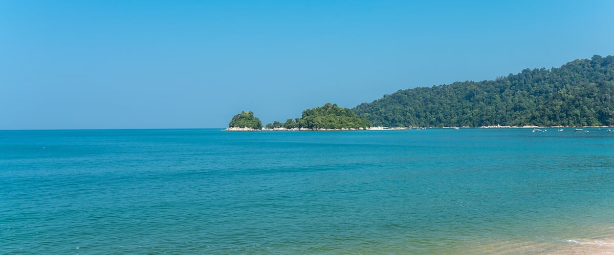 A beach with clear turquoise water, a sandy shore, and a forested hill in the background.