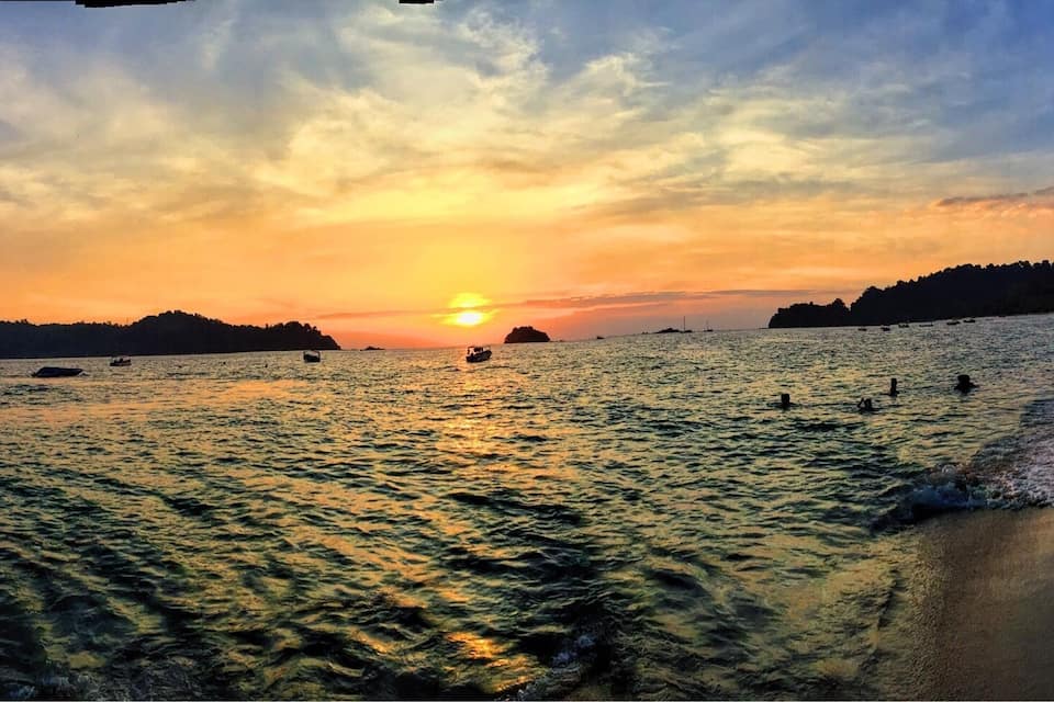 A beach at sunset with boats on the water and people on the shore.