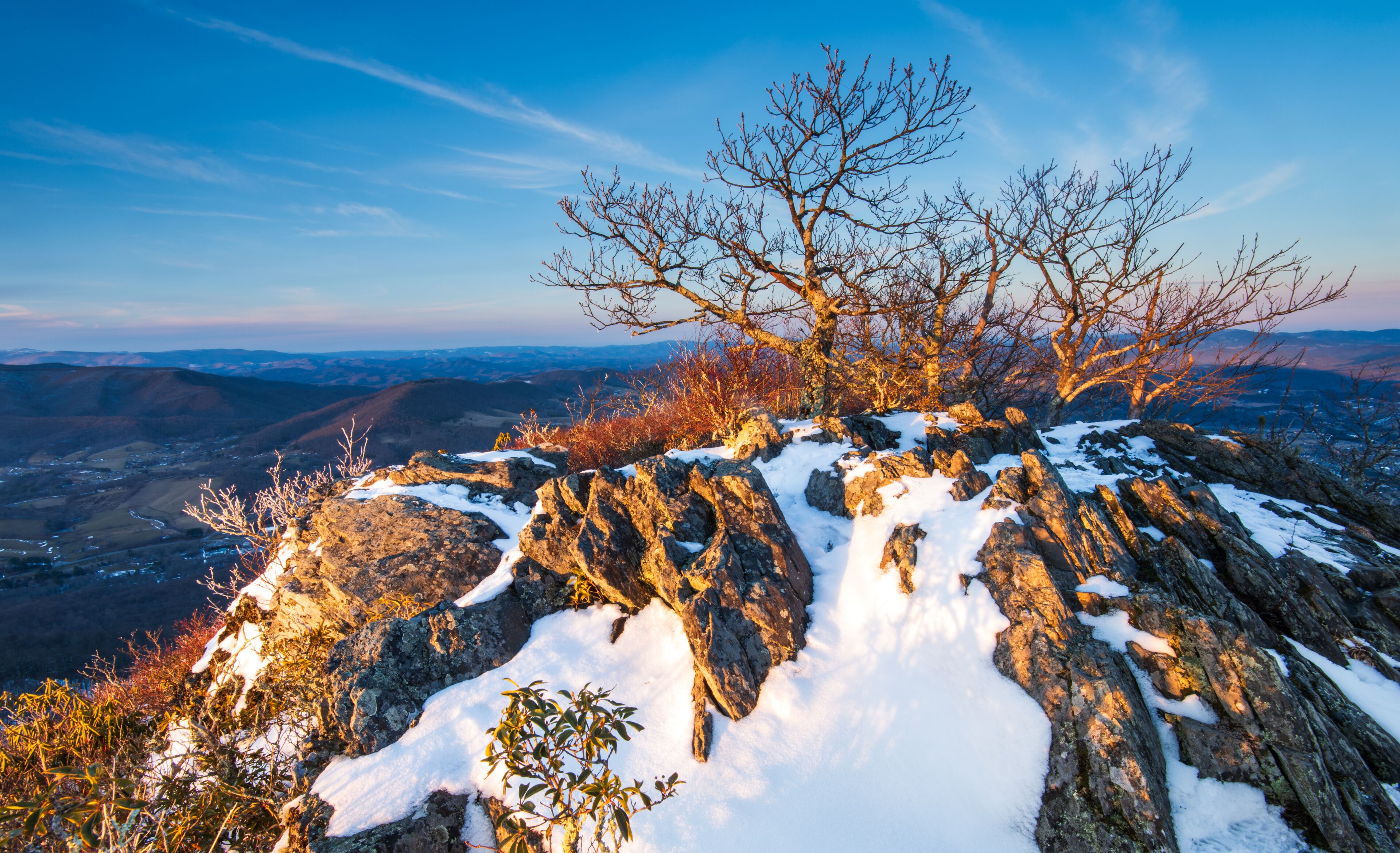 A snow-covered rocky mountain peak with a clear sky and distant valleys.