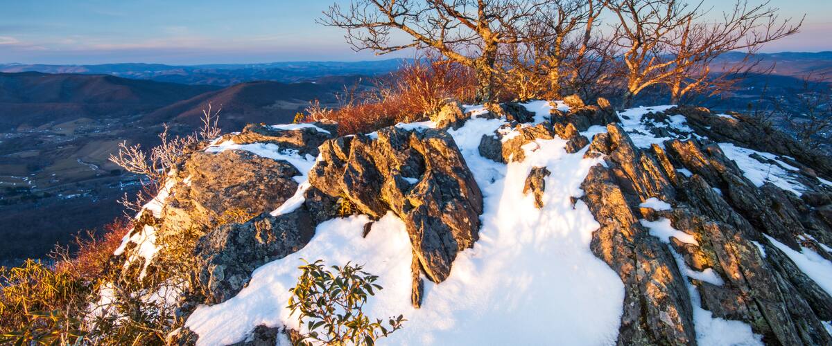 A snow-covered rocky mountain peak with a clear sky and distant valleys.