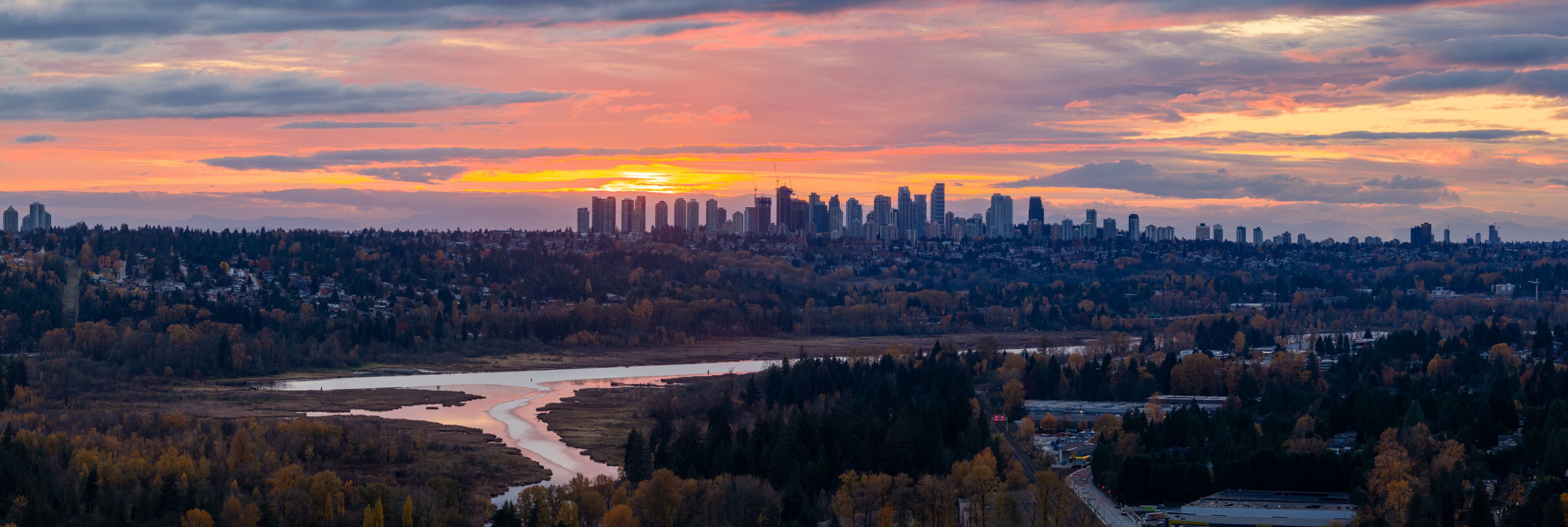 A city skyline at sunset with a river and forested area in the foreground.