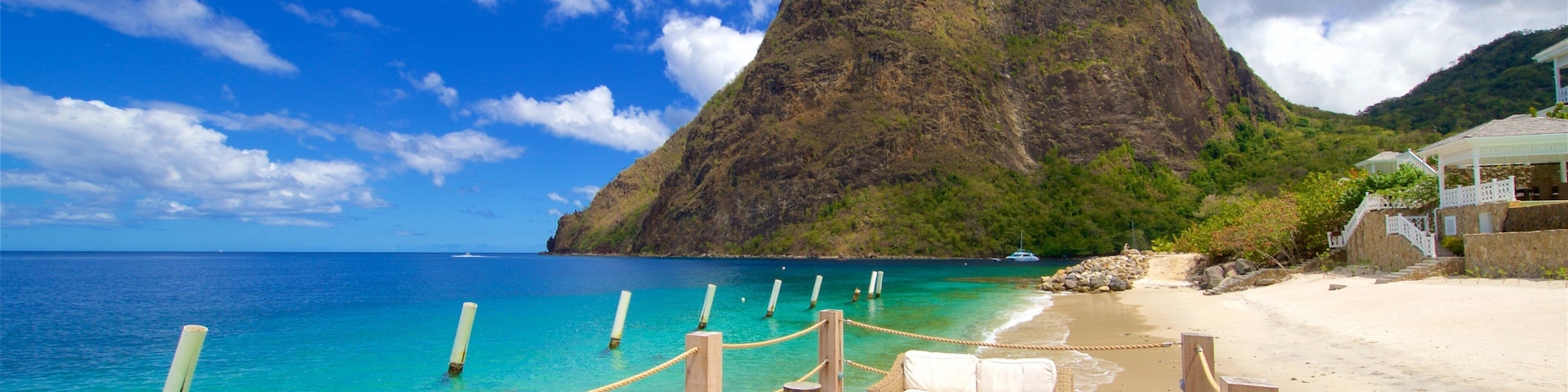 A wooden deck with wicker furniture overlooking turquoise waters, a sandy beach, and a prominent mountain in the background.
