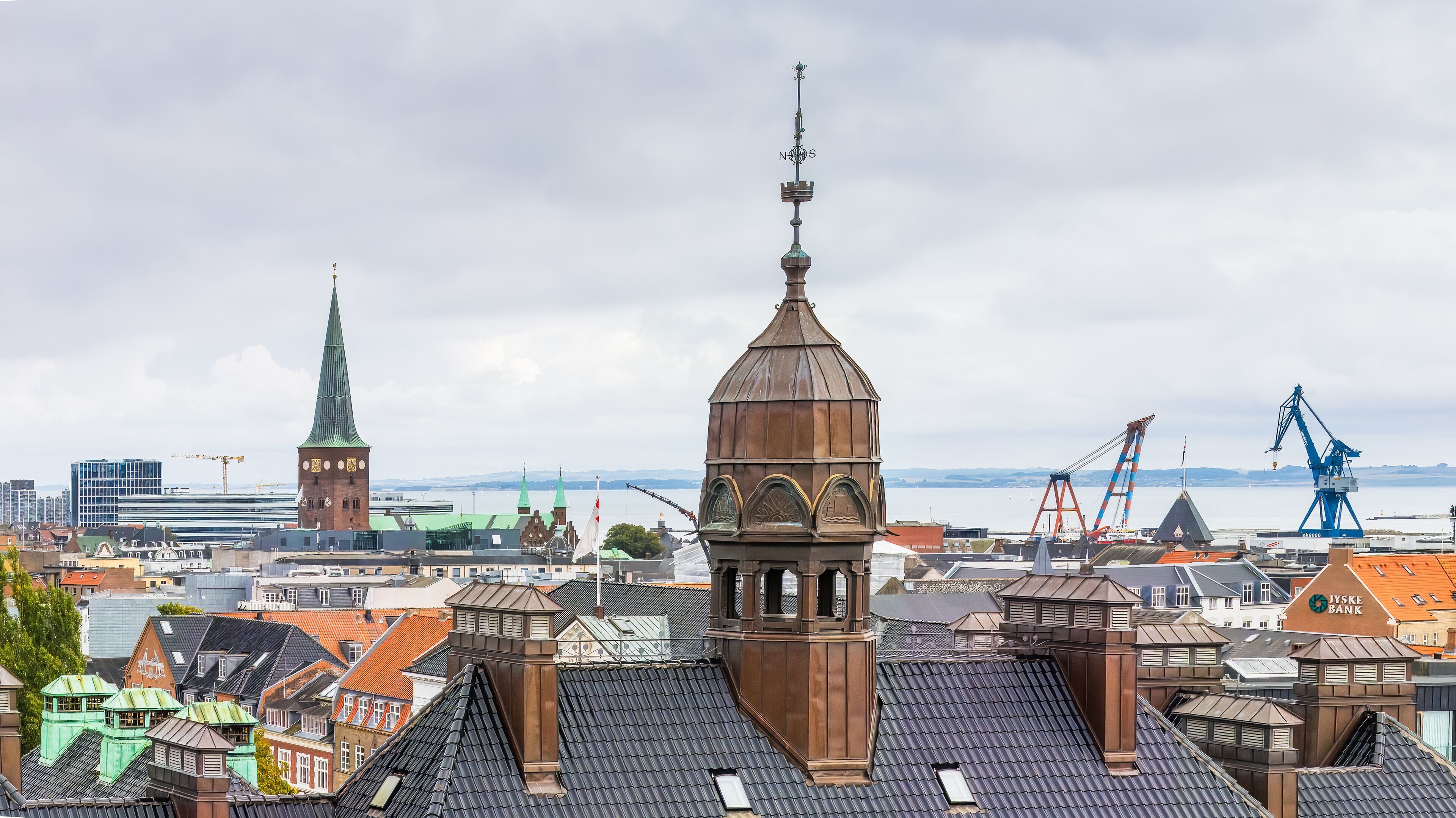 Skyline of Aarhus, Denmark, with the copper-topped towers and spire of historic buildings set against a backdrop of cranes, the waterfront, and a cloudy sky