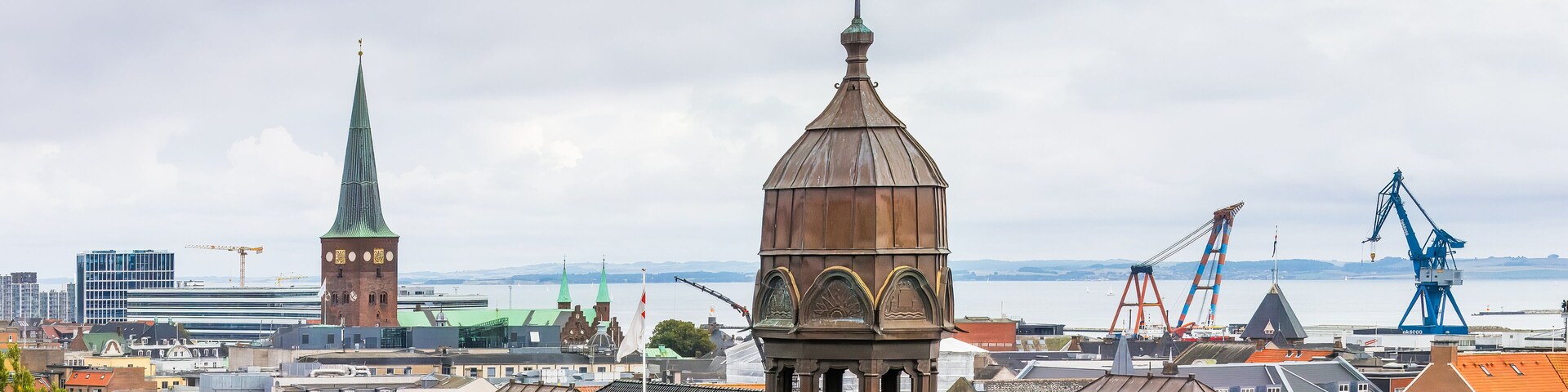 Skyline of Aarhus, Denmark, with the copper-topped towers and spire of historic buildings set against a backdrop of cranes, the waterfront, and a cloudy sky