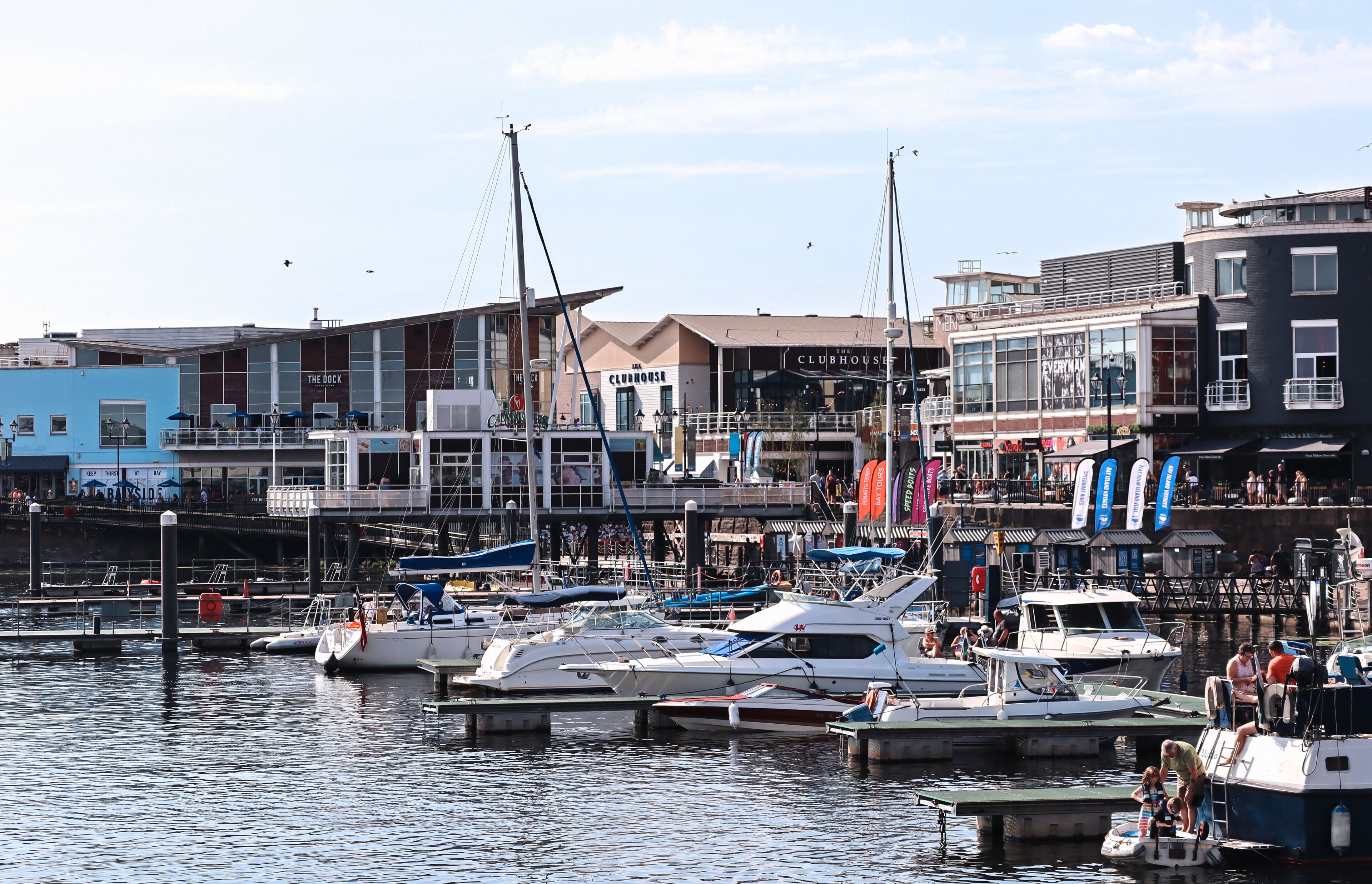 Uma marina com barcos atracados, um edifício com a placa 'The Clubhouse' e um calçadão à beira-mar.