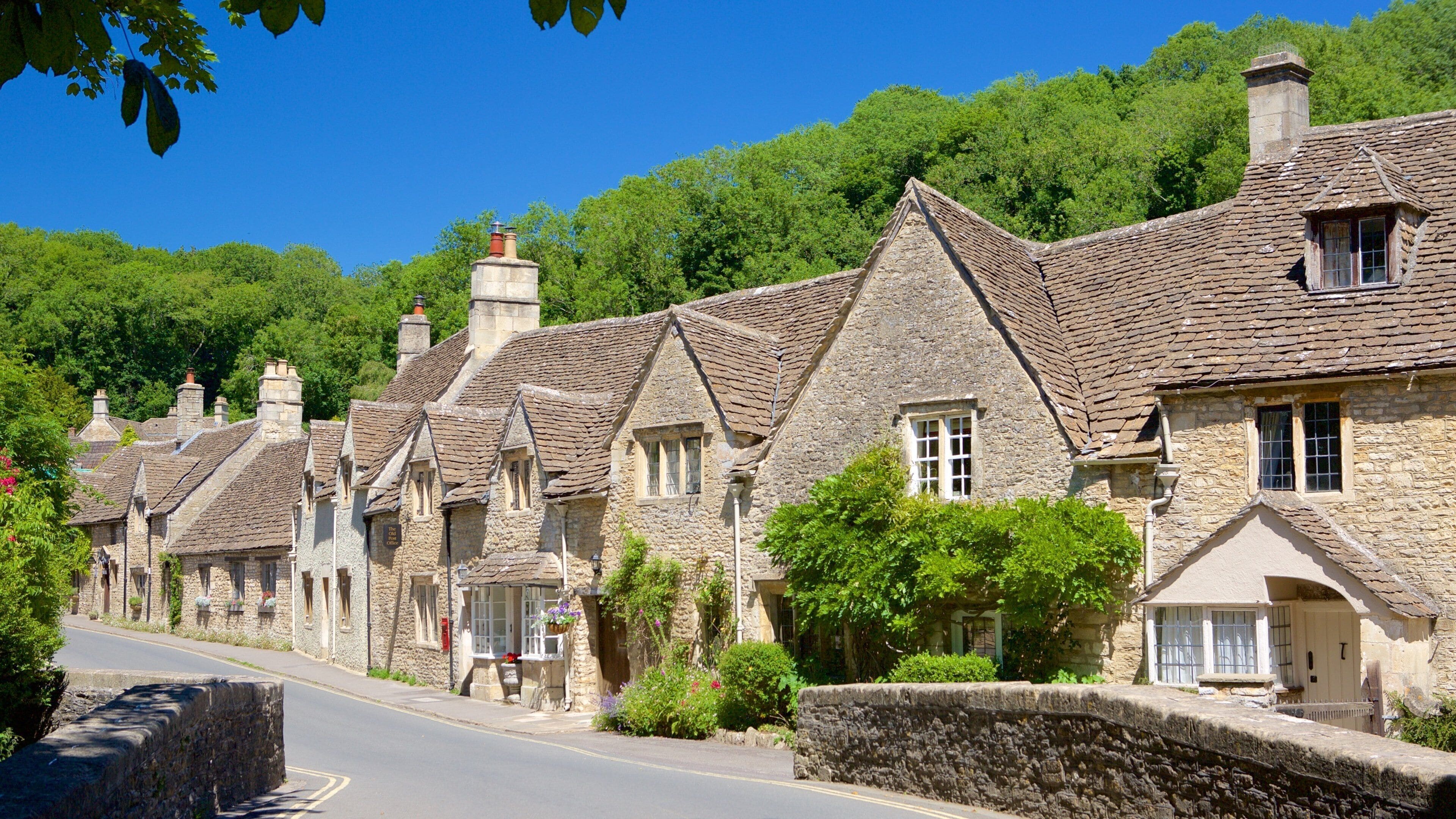 A row of stone houses with steeply pitched roofs, a stone wall, and lush greenery in the background.