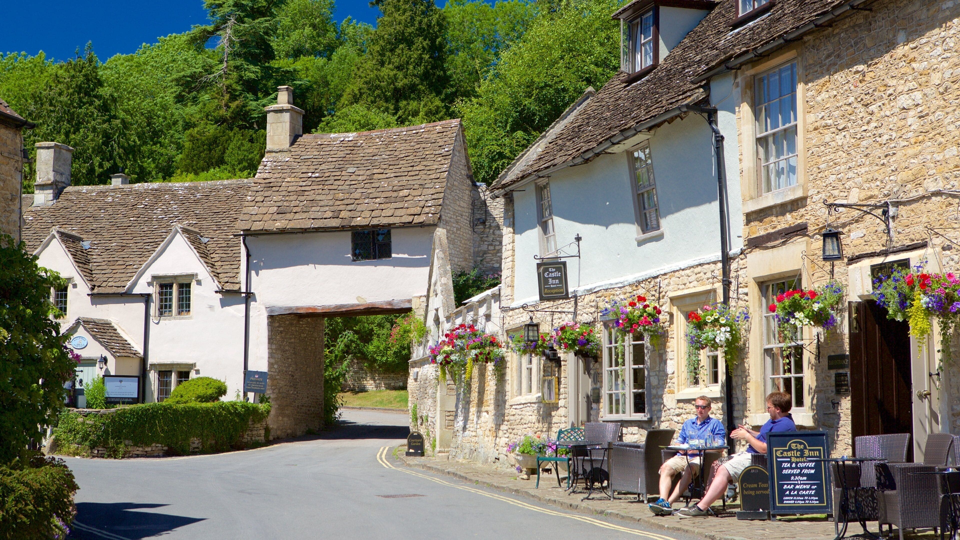 A quaint village street with traditional stone buildings, a pub named 'The Castle Inn', and outdoor seating with hanging flower baskets.