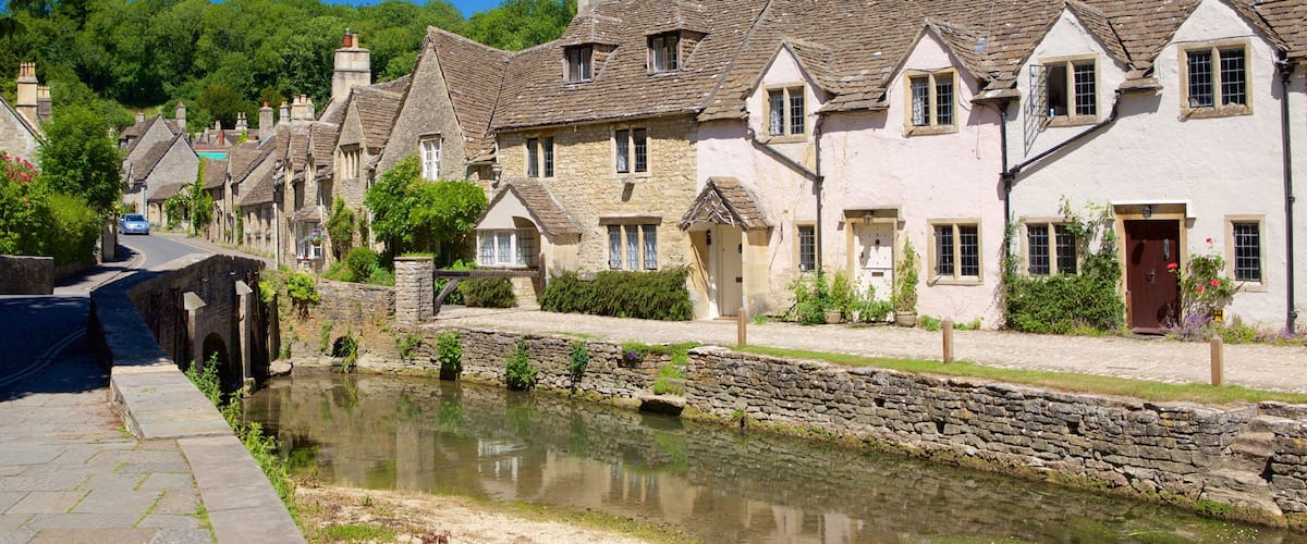 A row of stone houses with steeply pitched roofs, a narrow canal, and lush greenery in the background.