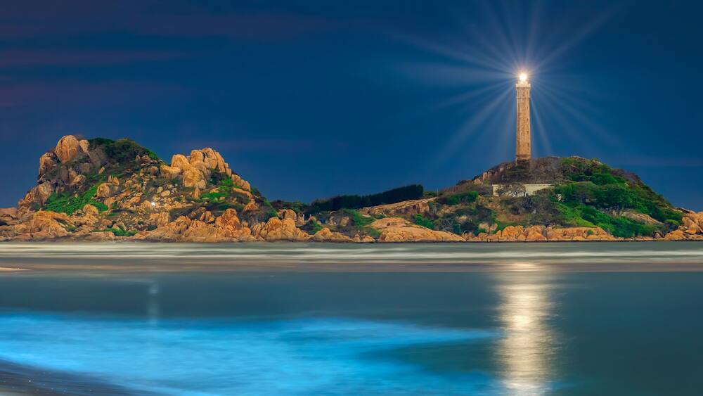 A lighthouse on a rocky island with a calm sea in the foreground.