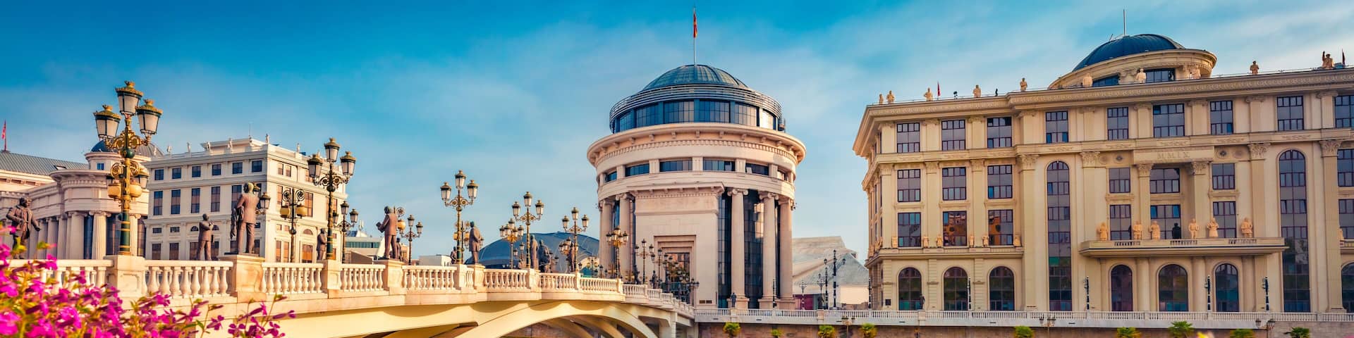 Blooming violet flowers on the shore of Vardar river. Exciting spring cityscape of capital of North Macedonia - Skopje with Archaeological Museum. Colorful view of Art Bridge.