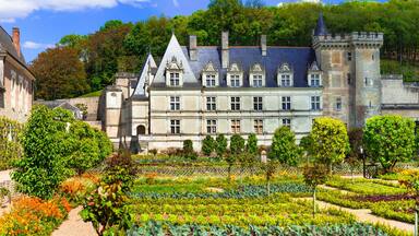 Ein historisches Schloss mit einem formalen Garten, umgeben von Bäumen und einer hügeligen Landschaft.