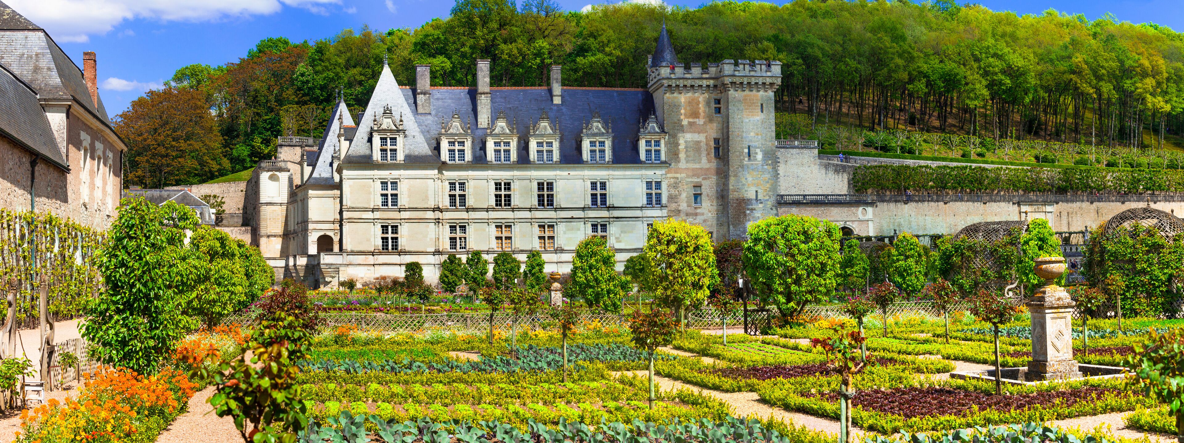 A historic château with a formal garden, surrounded by trees and a hilly landscape.