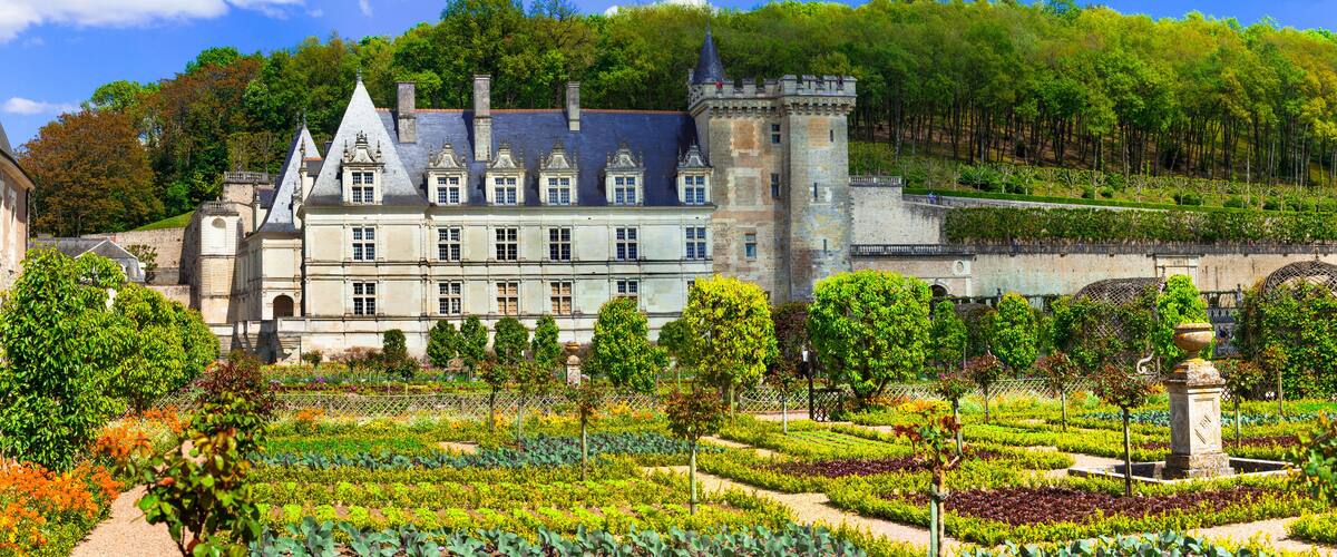 A historic château with a formal garden, surrounded by trees and a hilly landscape.