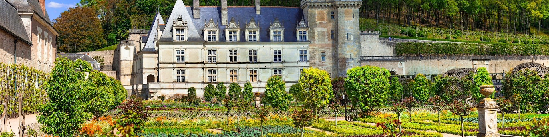 A historic château with a formal garden, surrounded by trees and a hilly landscape.