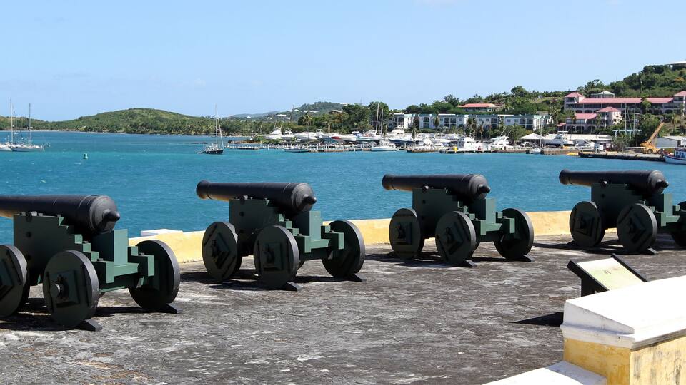 Tres cañones alineados en un paseo marítimo con un puerto deportivo y edificios al fondo.