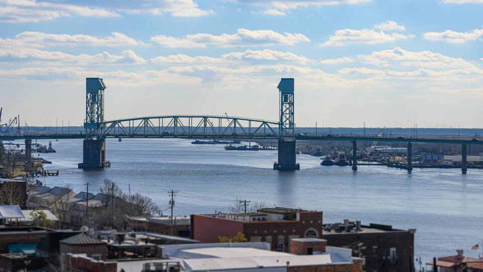 A large bridge spanning a river with industrial buildings and boats in the foreground.
