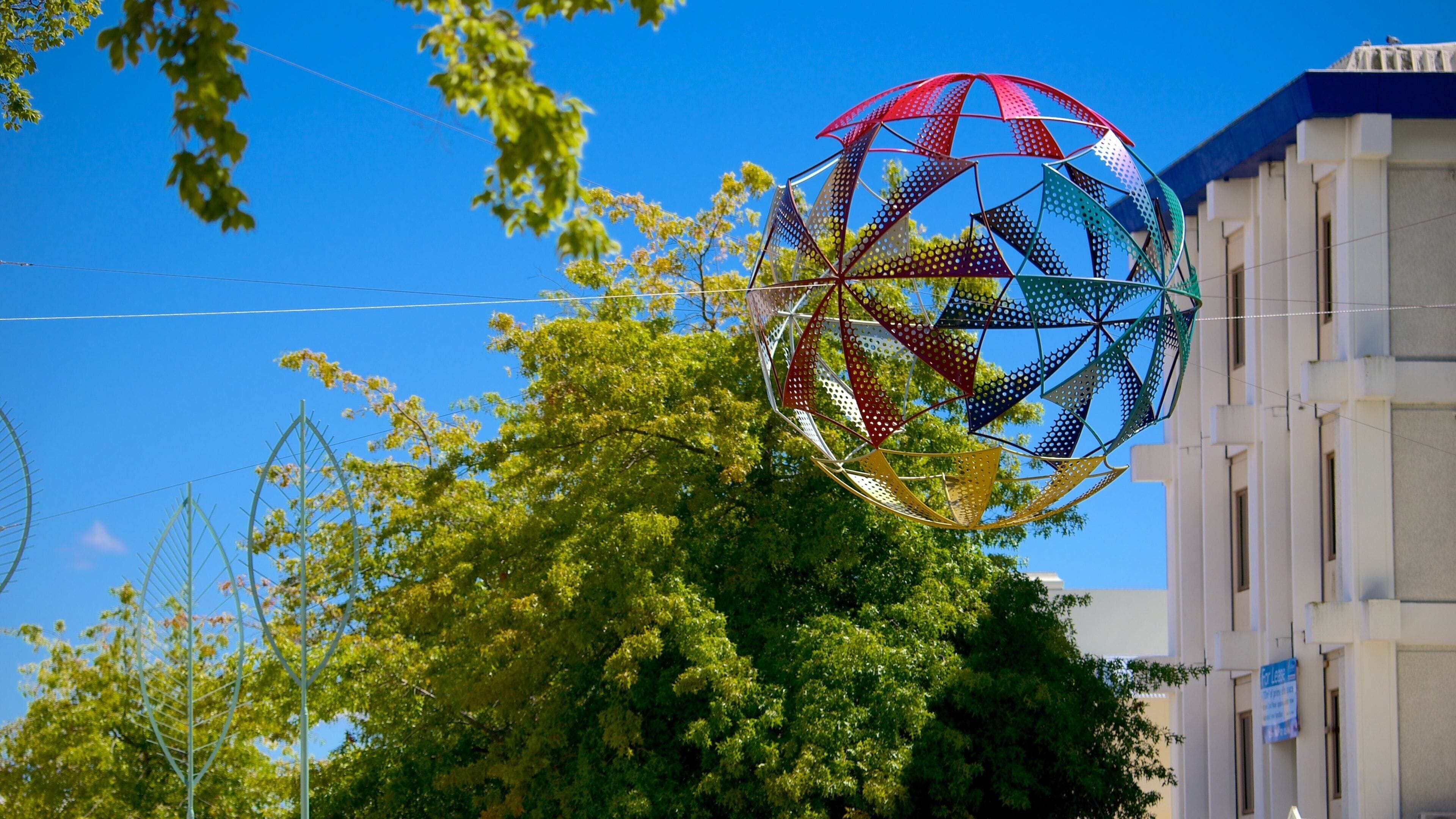A colorful spherical structure with a geometric pattern, set against a backdrop of a clear blue sky and green trees.