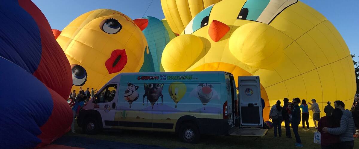 A group of large hot air balloons, including a yellow one with a face, and a white van with cartoon characters.