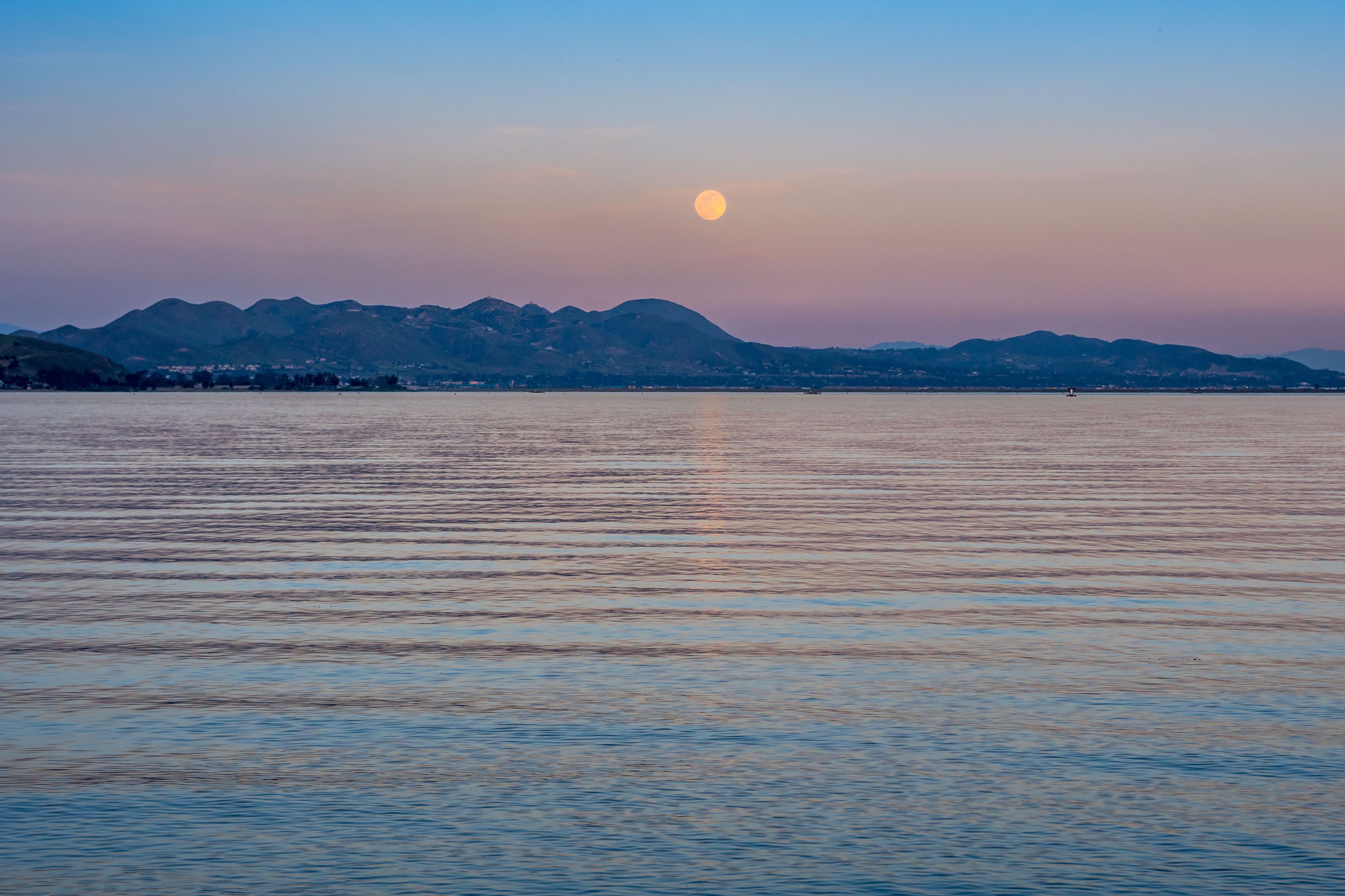 A calm lake with a mountain range in the background and a full moon in the sky.