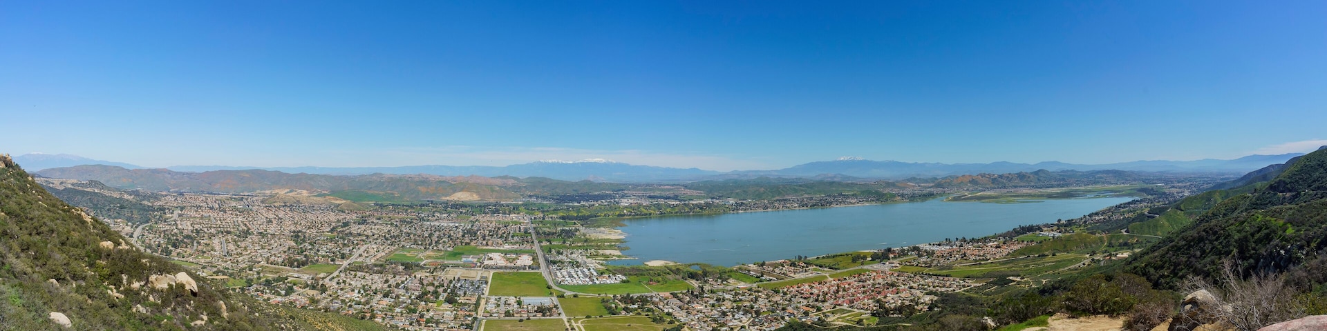A panoramic view of a coastal city with a large lake, surrounded by hills and mountains.