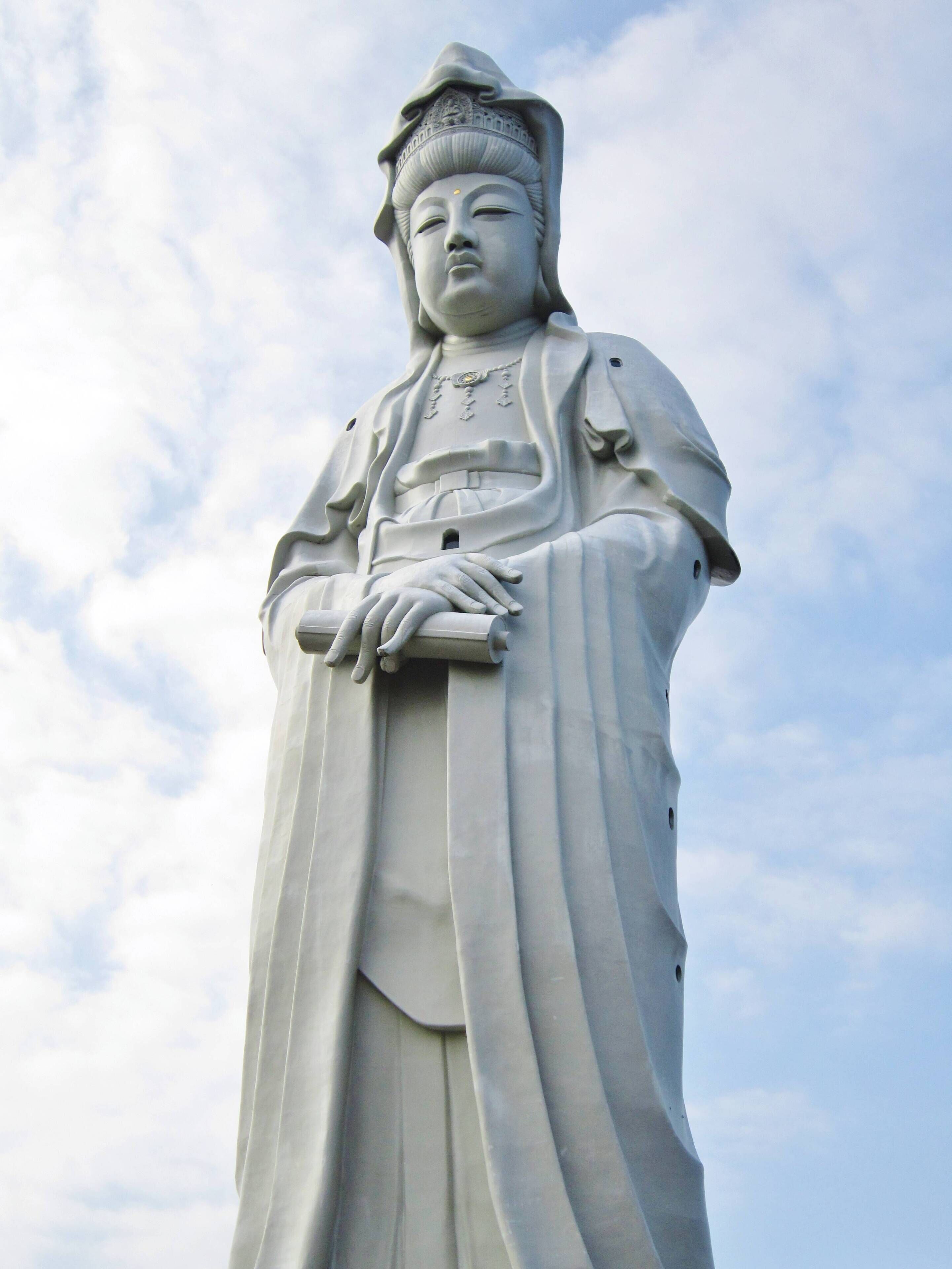 A large white statue of a figure in traditional attire with a draped robe and a headpiece, standing against a cloudy sky.