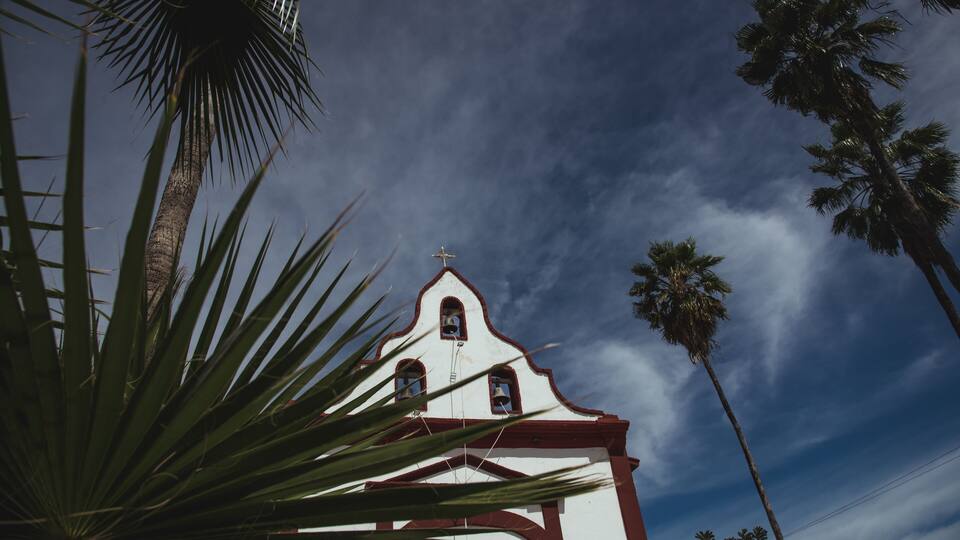 A white church with red trim and a cross on top, surrounded by palm trees.