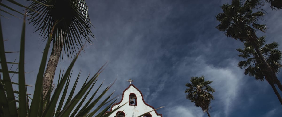 A white church with red trim and a cross on top, surrounded by palm trees.